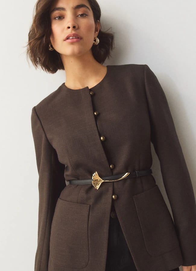 Woman wearing a dark brown, long-sleeved blazer with gold buttons and a thin black belt featuring a gold ginkgo leaf buckle.