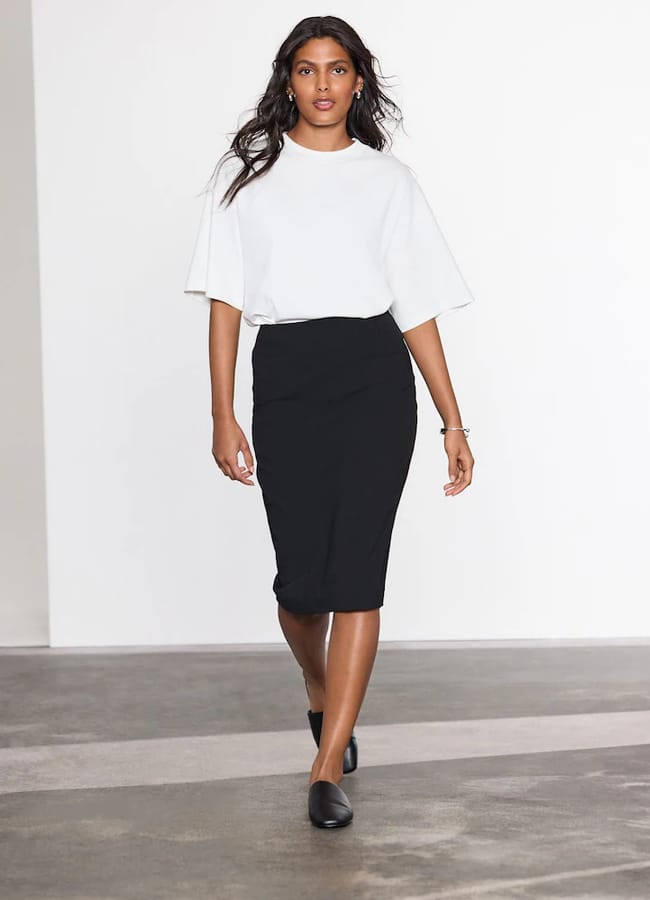 A woman wearing a white crew-neck T-shirt tucked into a black knee-length pencil skirt and black slip-on shoes.