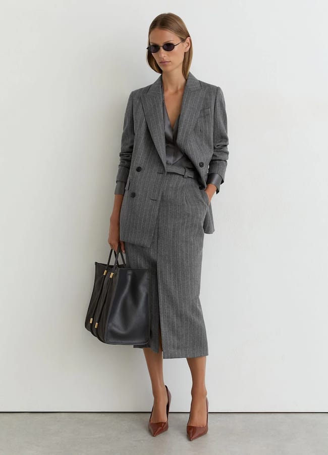 A model wearing a grey pinstriped blazer and matching midi pencil skirt, carrying a black tote bag and wearing brown heels and dark sunglasses.