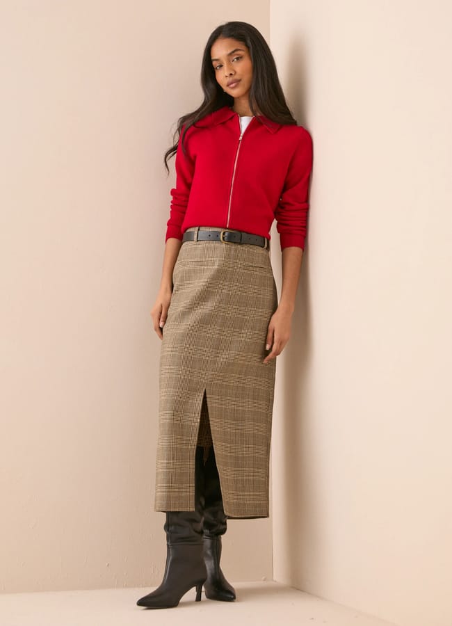 A woman wearing a red zip-up jumper, a checked pencil skirt with a front slit, and black boots.
