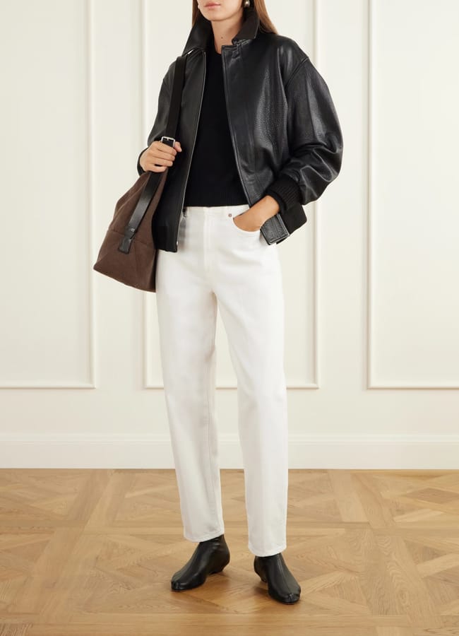 A person wearing one of the best women's leather jackets in a black bomber style, paired with a black jumper, white straight-leg jeans, and black leather ankle boots, whilst carrying a brown suede shoulder bag.