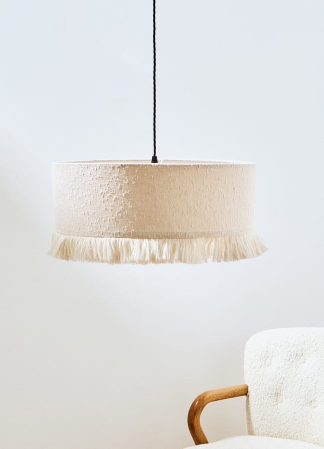Cylindrical pendant light with a textured off-white bouclé shade and fringe trim hanging from a black flex.