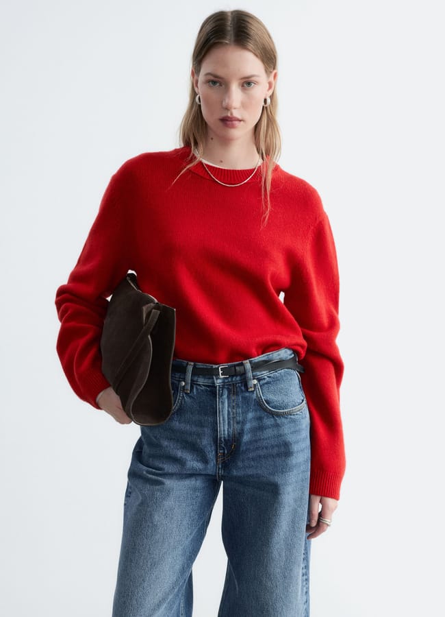 A person wearing a long-sleeved red jumper and blue jeans, holding a dark brown bag under their arm.