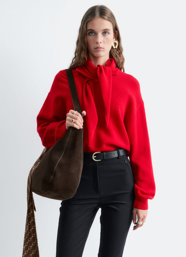 A woman wearing a red jumper with a scarf tie and black trousers carries a dark brown suede hobo bag with a patterned strap.