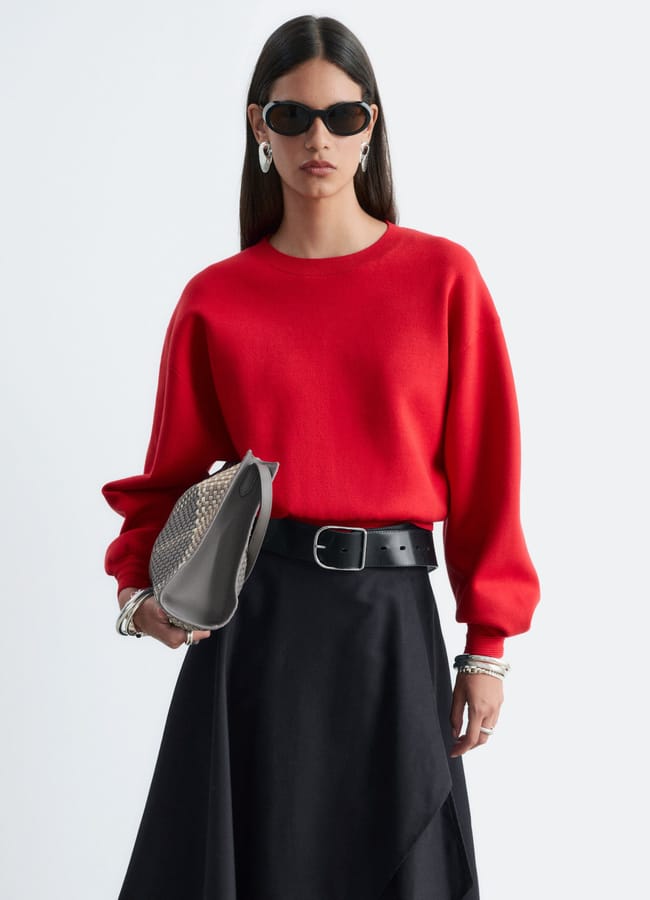 A person wearing a red jumper, black skirt with a black belt, and sunglasses, holding a grey textured handbag.