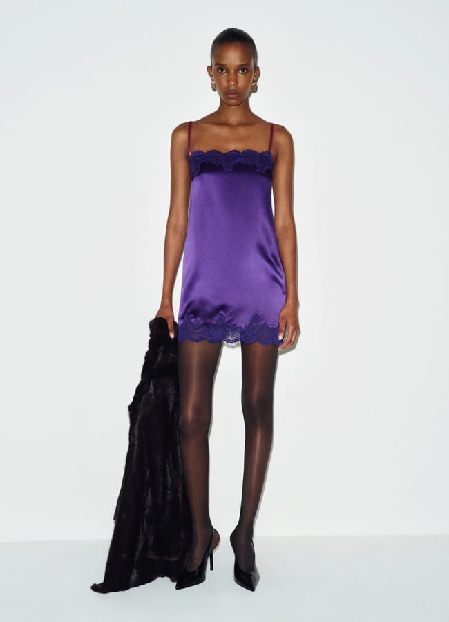 A person wearing a purple silk slip dress with lace trim, black tights, and black high heels, holding a dark coat.