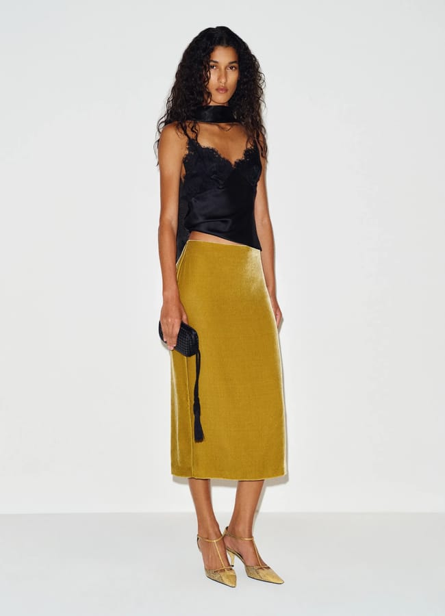 A person wearing a black lace camisole, a mustard yellow midi pencil skirt, and metallic t-bar heels, holding a small black clutch.