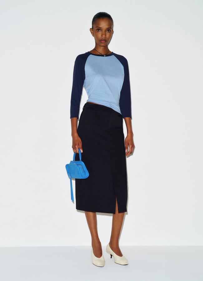 A model wears a light blue and navy baseball-style top, a black midi pencil skirt with a front slit, and cream-coloured heels, whilst holding a small blue handbag.