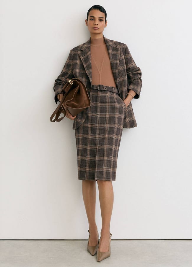 A model wears a brown checked blazer and matching belted pencil skirt over a tan polo-neck top, holding a brown leather handbag and wearing tan heels.