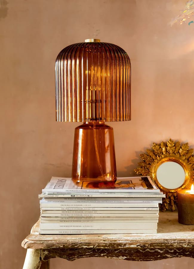 Amber-coloured ribbed glass Rockett St George table lamp sitting on a stack of magazines next to a small gold sunburst mirror and a candle.