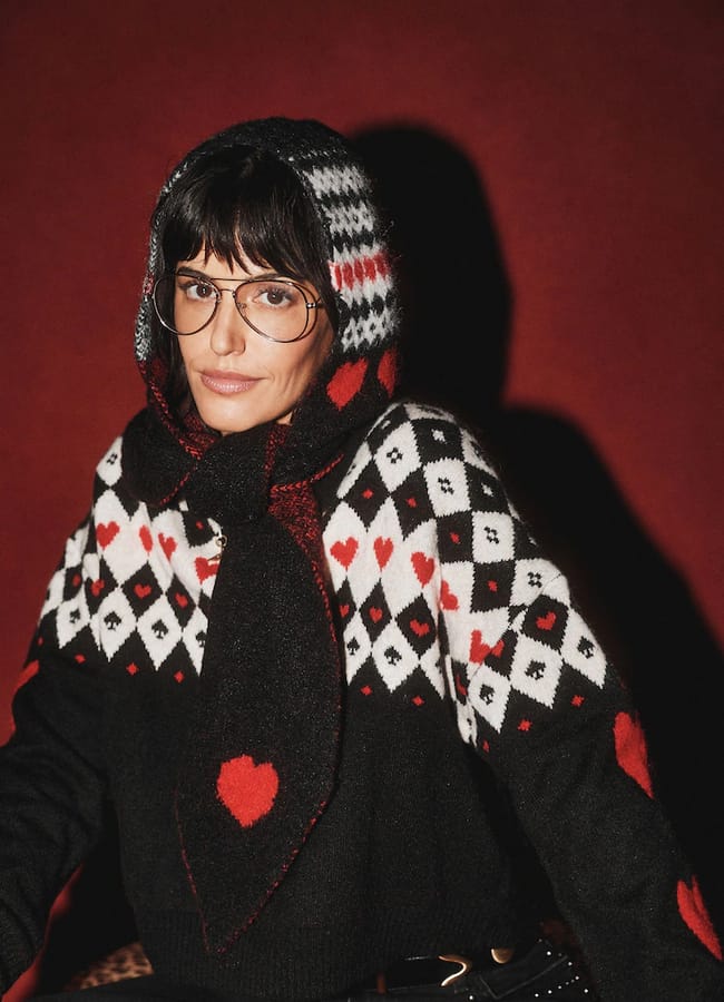A person wearing glasses and a black Rockett St George hooded jumper patterned with white and red diamonds and hearts.