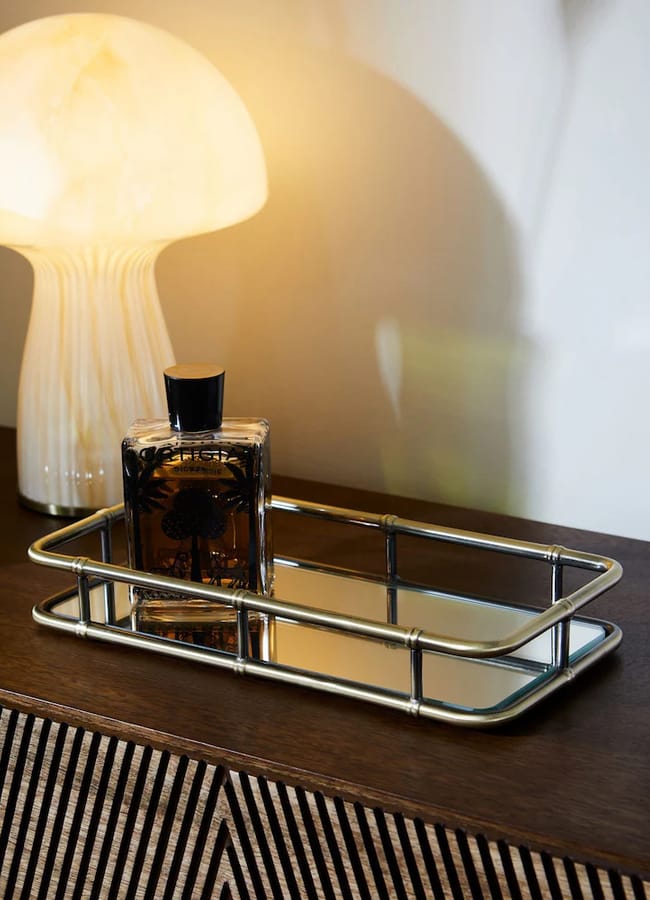 A Rockett St George rectangular mirrored tray with a metal frame sits on a wooden surface next to a glass perfume bottle and a mushroom-shaped table lamp.