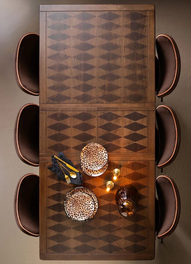 Overhead view of a Rockett St George rectangular wooden dining table with a diamond pattern, set with two stacks of leopard-print plates, gold cutlery, and two small candles, flanked by six brown chairs.
