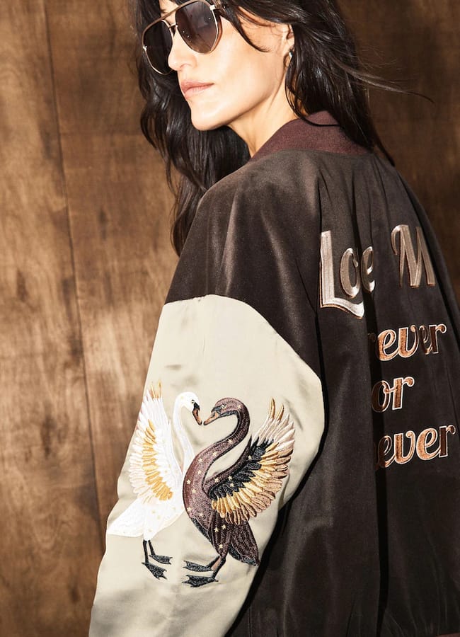 A Rockett St George person wearing a brown and tan bomber jacket with embroidered swans on the sleeve and text on the back.