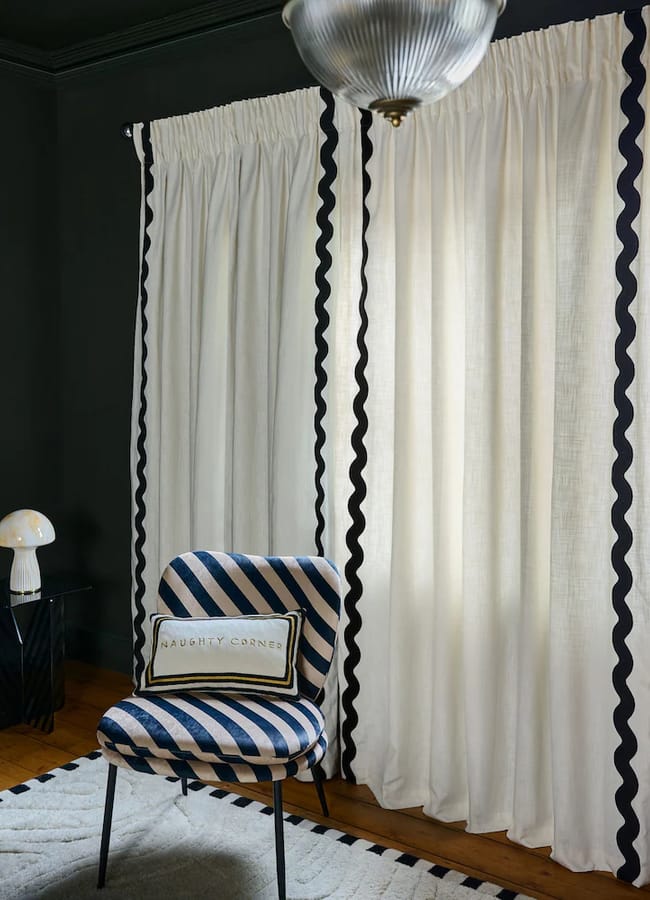Off-white Rockett St George curtains with black wavy trim hang in front of a blue and white striped chair with a rectangular cushion embroidered with the words Naughty Corner.