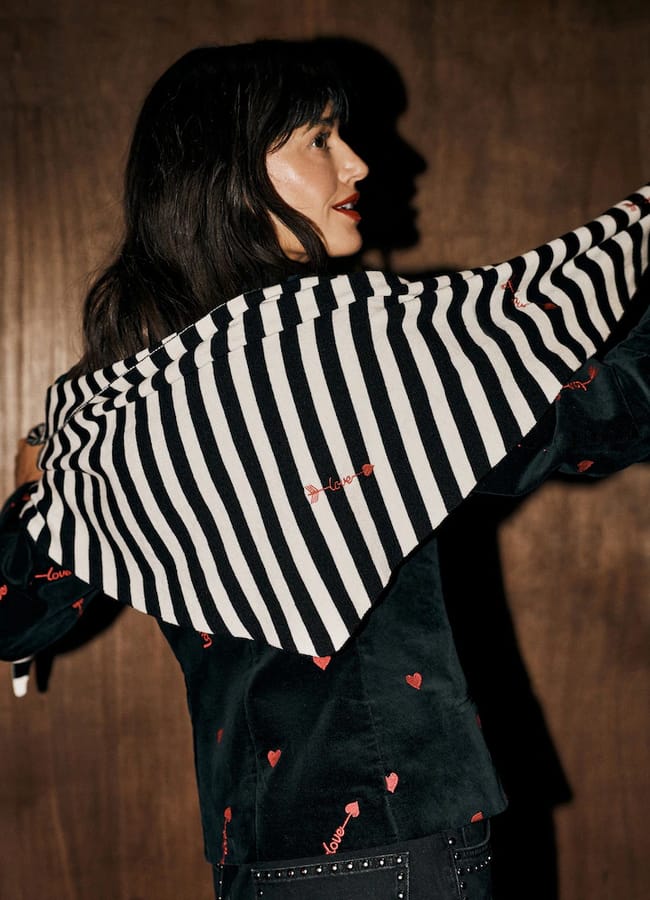 A Rockett St George woman wears a black velvet jacket with red heart and love embroidery over a black and white striped scarf.