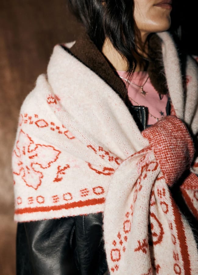 A person in a pink shirt, black leather jacket, and a cream-coloured Rockett St George shawl featuring a red floral pattern.