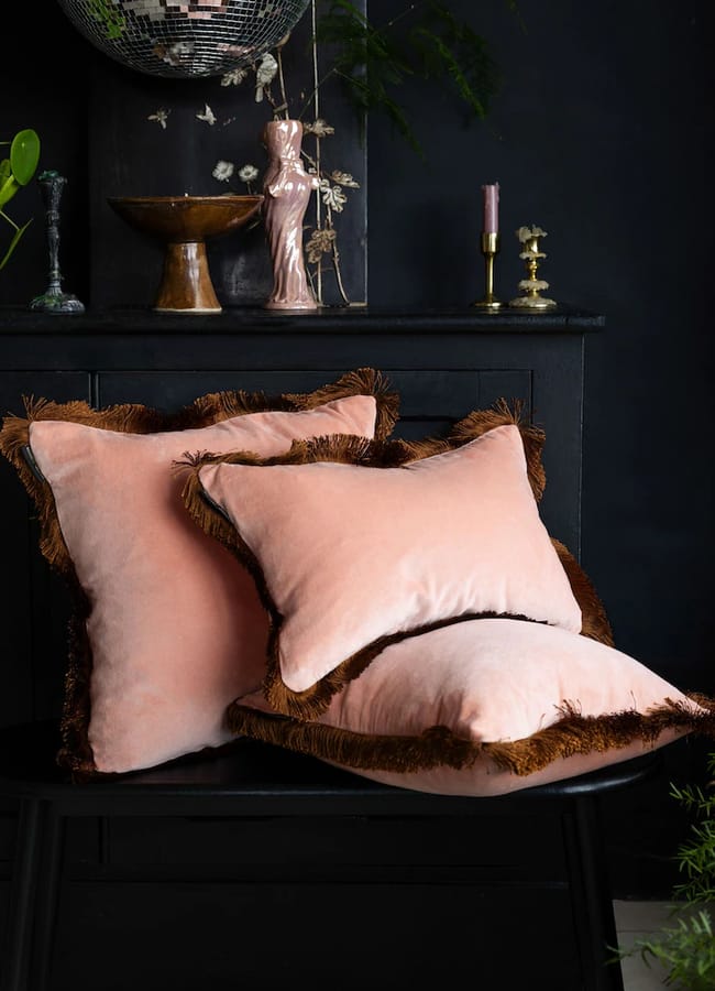 Three light pink Rockett St George throw cushions with dark brown fringe trim stacked on a black bench.