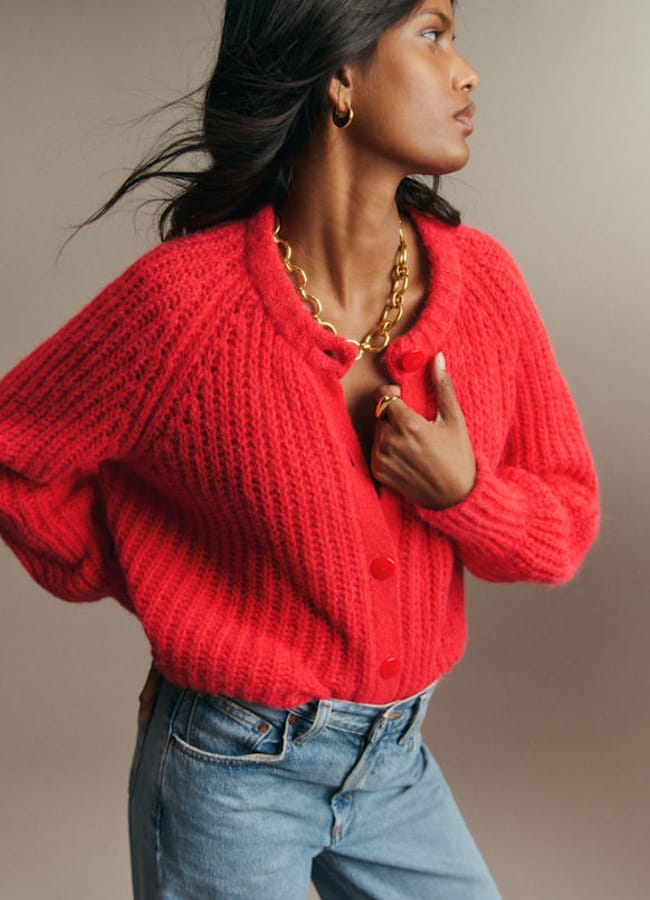 Person wearing a bright red knitted button-up cardigan and blue jeans, accessorised with a gold chain necklace and hoop earrings.