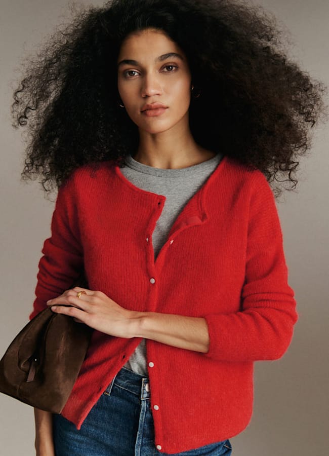 A person wearing a bright red knitted button-up cardigan over a grey T-shirt and blue jeans, holding a brown suede bag.