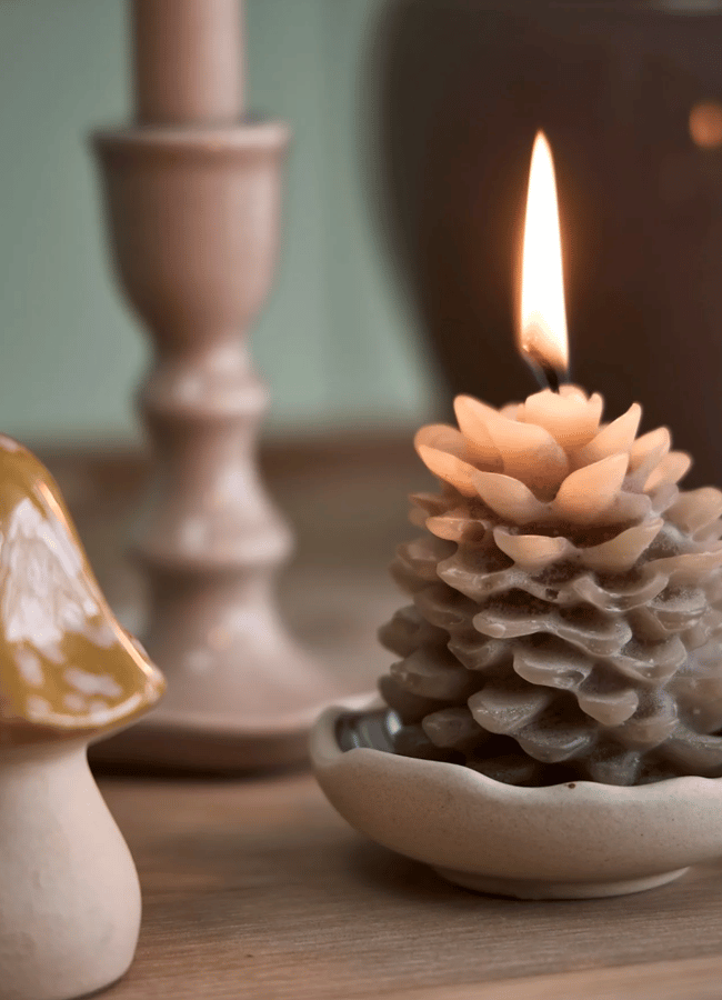 A lit pinecone-shaped candle sits in a small dish next to a decorative mushroom ornament and a candlestick, perfect for autumn tablescape ideas.