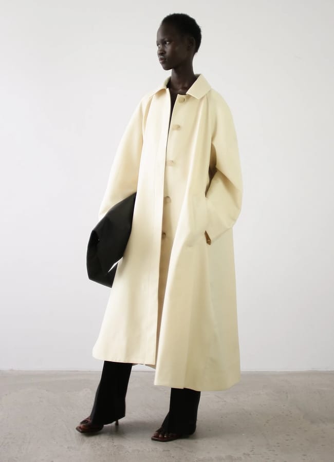 A person wears a long, cream-coloured coat over dark trousers, carrying a large black clutch bag from a black owned fashion brand.