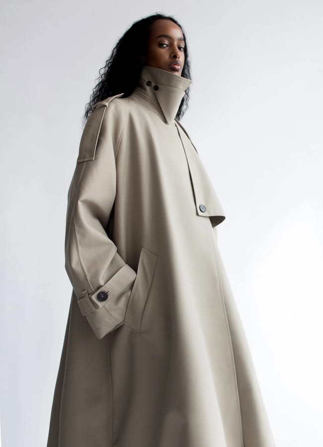 A person wearing a beige, oversized trench coat from a black-owned fashion brand with a high collar.