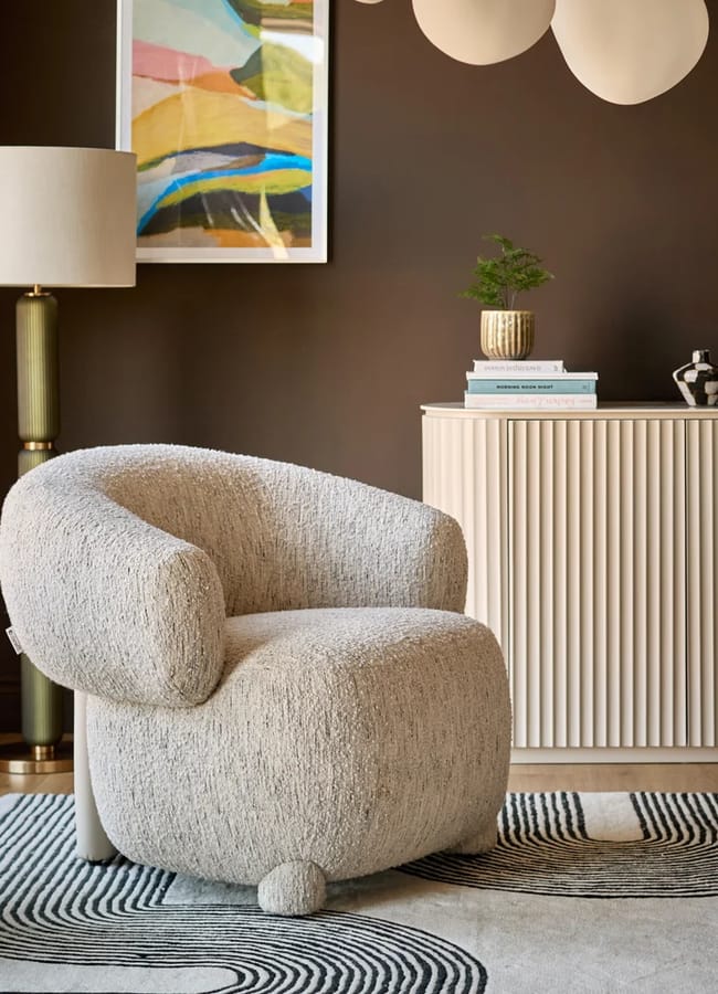 A rounded, textured cream-coloured bouclé accent chair sits on a rug with a black-and-white line pattern in front of a fluted sideboard.