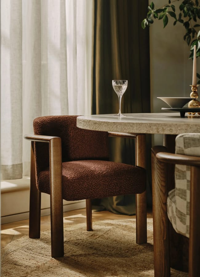 A round stone dining table with a wine glass and candlestick, flanked by two bouclé upholstered armchairs on a woven rug.