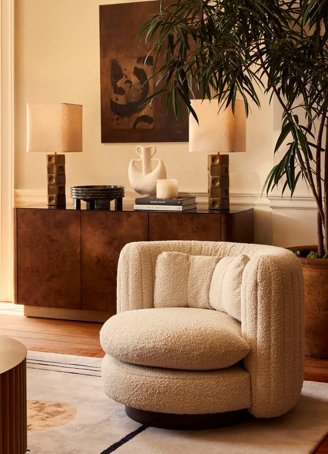 A cream-coloured, curved bouclé swivel armchair sits on a rug in front of a wooden sideboard topped with two lamps, a white vase, and stacked books.