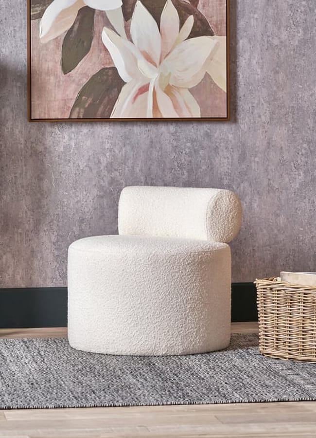 A white, barrel-shaped bouclé armchair with a low back, positioned on a grey rug next to a tall lamp and a wicker basket.