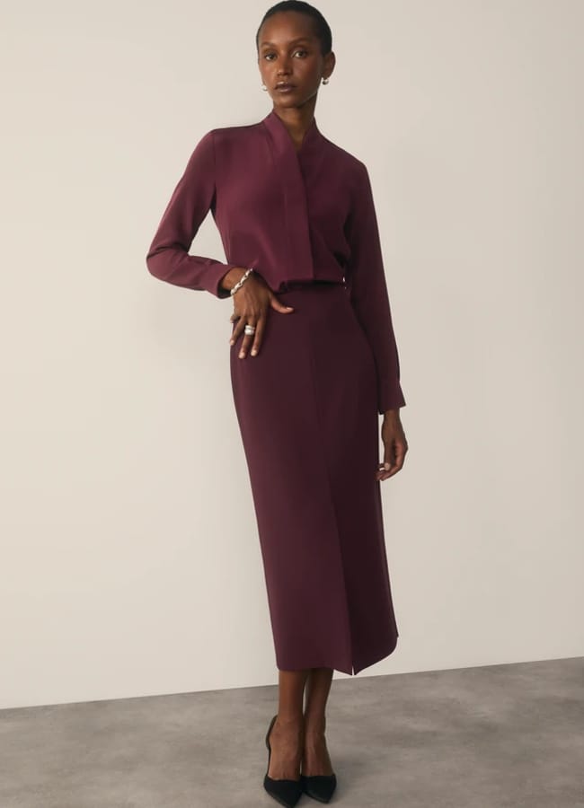 A woman wears a long-sleeved, deep plum blouse with a high neckline tucked into a matching midi-length pencil skirt, paired with black pointed-toe court shoes.