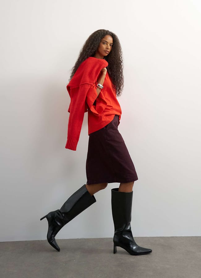 A person wearing a red jumper draped over their shoulders, a burgundy pencil skirt, and black knee-high boots walks against a white background.