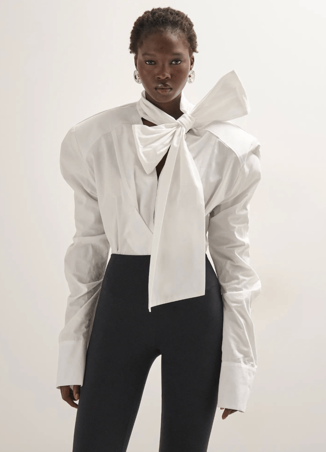 A model wears a white long-sleeved blouse from a black-owned fashion brand with dramatic puffed shoulders and a large bow at the neck, paired with black fitted trousers.