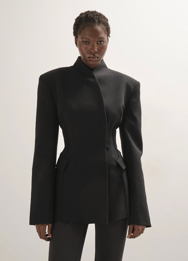 A model wearing a black structured blazer from a black-owned fashion brand, featuring a high collar and a cinched, hourglass waist.