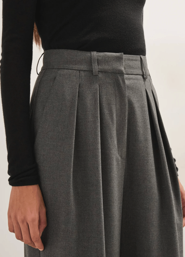 A person wears a black top from a black owned fashion brand tucked into high-waisted, pleated grey trousers with belt loops.