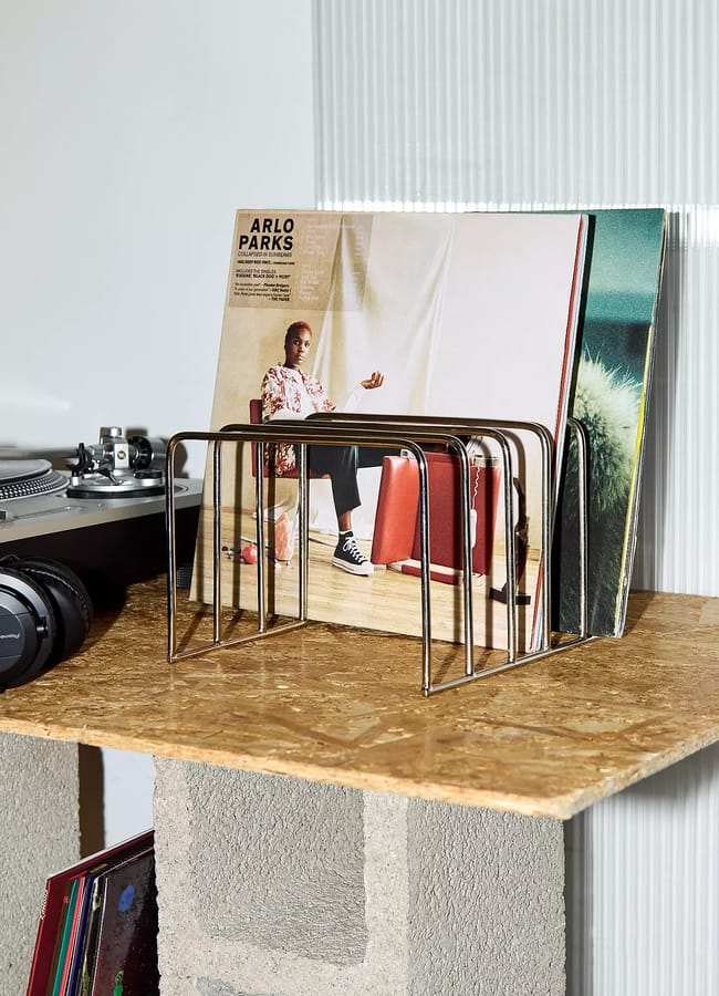 A wire record rack on an OSB shelf holds several vinyl albums, including Arlo Parks’ Collapsed in Sunbeams, as part of a curated listening room interior.