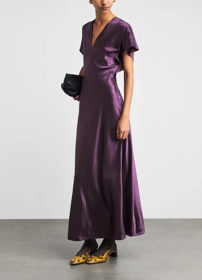 A person wears a floor-length, deep purple v-neck dress, carrying a black clutch and wearing leopard-print block-heel shoes.