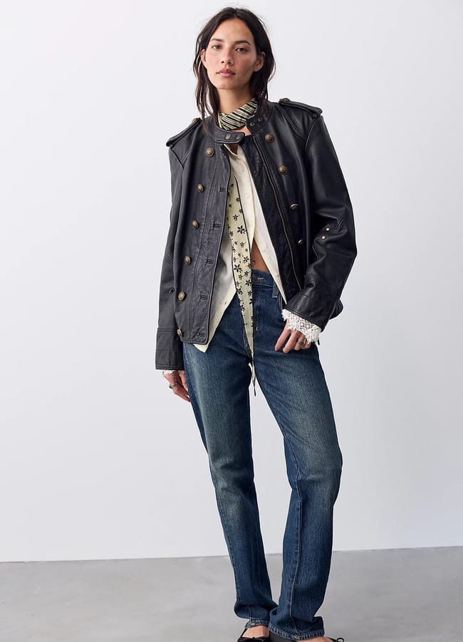 A woman wears one of the best women’s leather jackets in a black military style over a cream-coloured floral blouse and straight-leg blue jeans.
