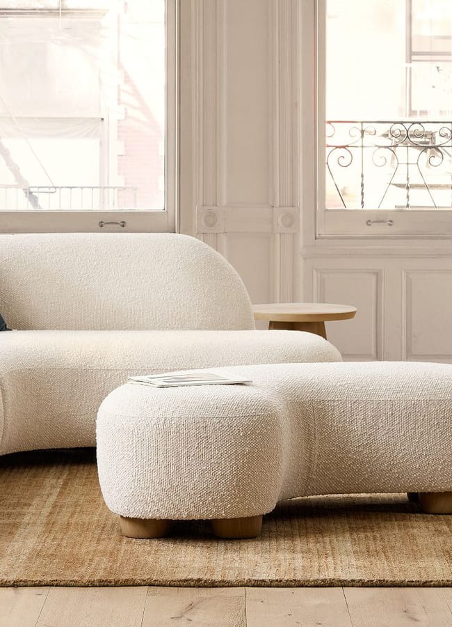 Curved cream-coloured bouclé sectional sofa and matching ottoman on a woven area rug in front of a window.