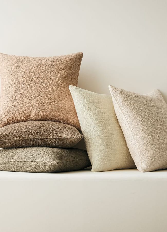 Three textured bouclé scatter cushions stacked on the left and two textured scatter cushions standing upright on the right, in shades of tan, brown, and cream.