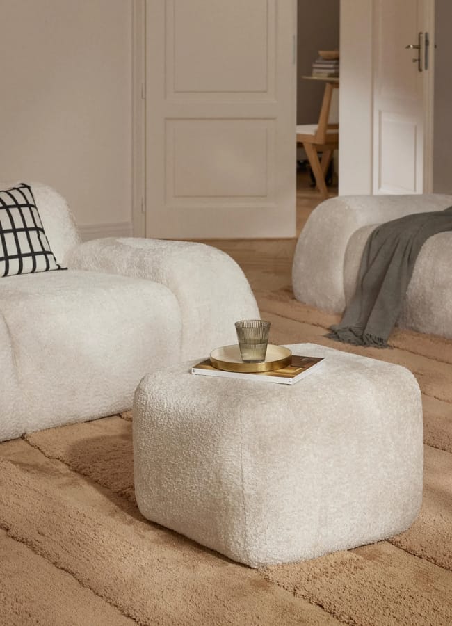 Two white textured bouclé armchairs and a matching ottoman arranged on a tan rug, with a glass and tray on the ottoman.