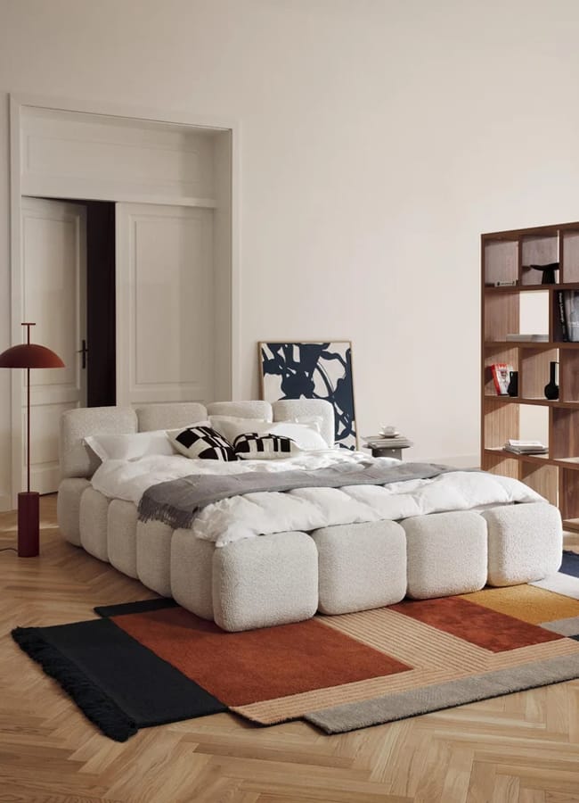 A low, white, bouclé block-shaped bed sits in a bedroom next to a floor lamp, a wooden shelving unit, and a patterned area rug.