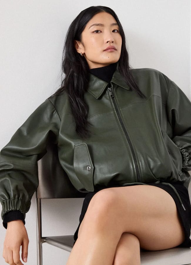 A person wearing an olive green leather bomber jacket, one of the best women's leather jackets, over a black polo neck and skirt, sitting on a chair.
