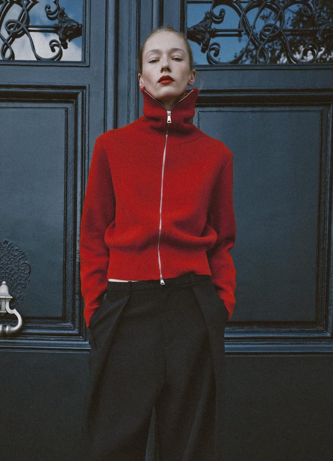 A person in a red, high-collar zip-up jumper and black trousers stands in front of a dark, ornate double door.