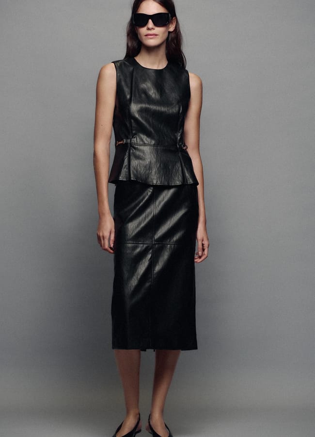 A person wears a black sleeveless peplum top and a matching midi pencil skirt with black sunglasses and pointed-toe flats.